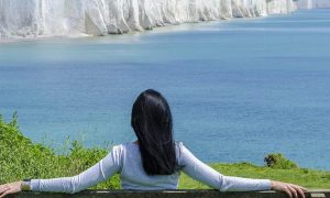 woman facing backward sits on bench looking at blue ocean