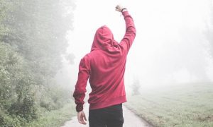 man wearing red hoodie facing backward walks along footpath cheer up between bush green grass in foggy weather