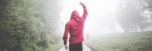 man wearing red hoodie facing backward walks along footpath cheer up between bush green grass in foggy weather