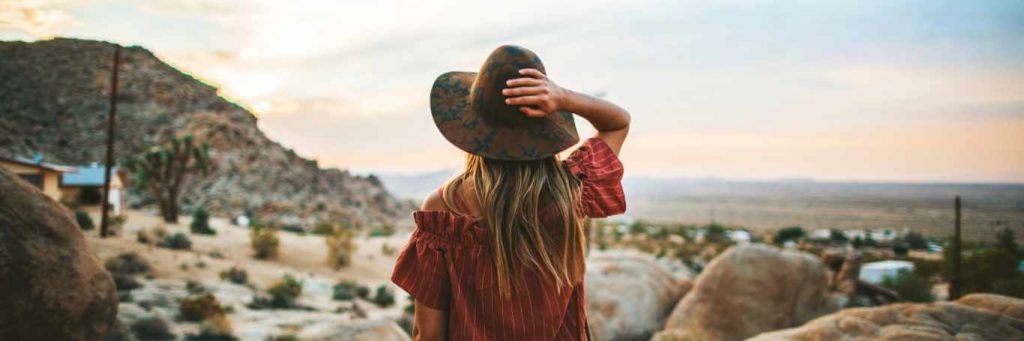 long blonde hair woman hand on hat stands on rock looking at desert mountain in blue cloudy sky