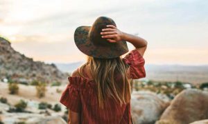 long blonde hair woman hand on hat stands on rock looking at desert mountain in blue cloudy sky