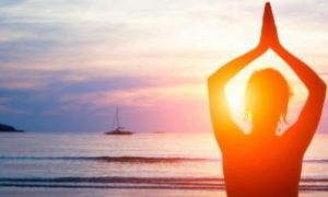 woman facing backward meditates on beach in blue sunny sky