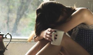 woman sits alone head on knees holding white mug feeling sad tired exhausted