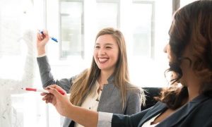 two business women stands in office happily discussing writing on whiteboard