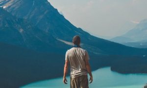 man stands beside lake looking at mountain in foggy weather