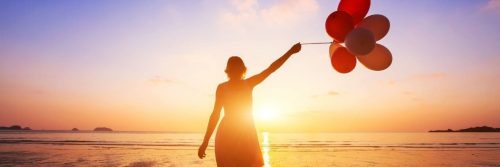 woman stands facing backward on beach holding balloons watching sunset