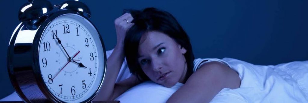 woman lies on bed scratches hair looks at clock at late night