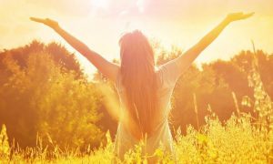 woman stands facing backward raising hands gratitude life in field in sunny sky