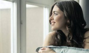 young woman sits beside window smiling hugging white pillow