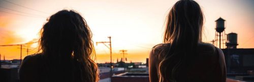 two women stands facing backward looking at inner city street sunset sky
