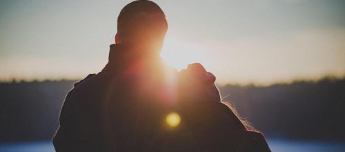 couple stands watching sunset while girl lying her head on boyfriend shoulder
