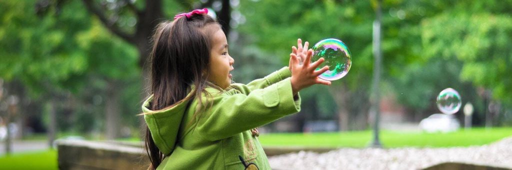 little girl stands in park playing with bubble