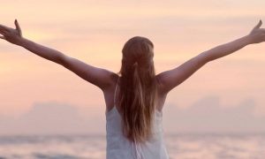 woman stands facing backward beside beach raising hands gratitude life in sunset sky