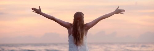 woman stands facing backward beside beach raising hands gratitude life in sunset sky