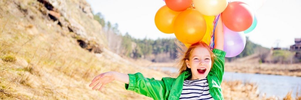 girl stands on field beside lake holding balloons happily smiling