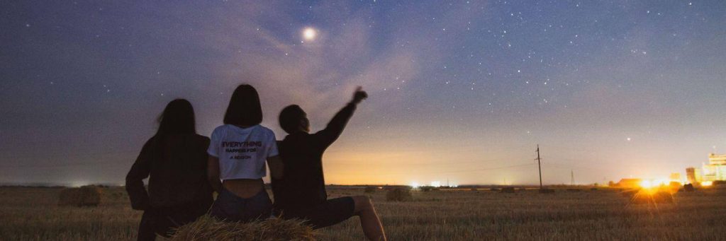 three people sit on grass facing backward watching stars while man pointing at blue sky moon