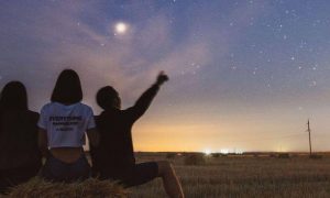 three people sit on grass facing backward watching stars while man pointing at blue sky moon