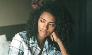 curly black hair woman sits hand on cheek in bedroom thinking
