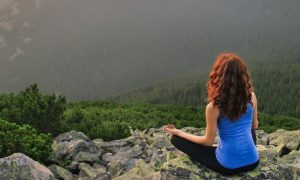 woman sits on mountain top focuses on meditation facing forest