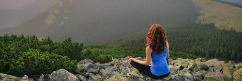 woman sits on mountain top focuses on meditation facing forest