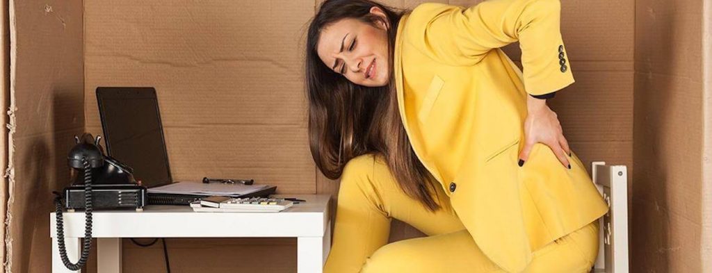 woman wearing yellow suit sits on low chair feeling lower back hurt while working on laptop