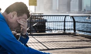 man sits on bench near seaport hand in face feeling sad