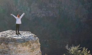 woman stands on rock raising hand feeling grateful of life