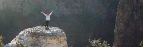 woman stands on rock raising hand feeling grateful of life