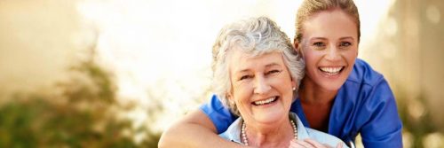 grandmother sits happily smiling beside granddaughter standing hugging from behind