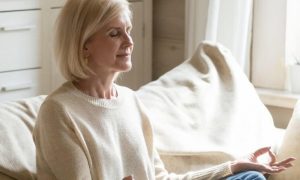 elderly woman sits on couch focuses on meditation breathing in living room