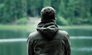 man stands alone facing backward looking at lake mountain