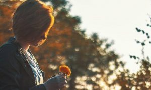 short hair woman holds small orange flower standing in forest in sunny sky