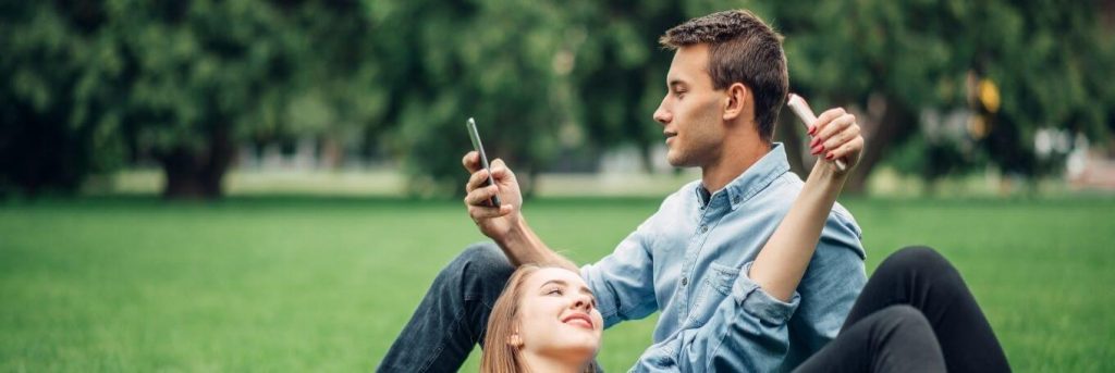 man sits on green grass using mobile phone while girlfriend lying on his leg taking selfie