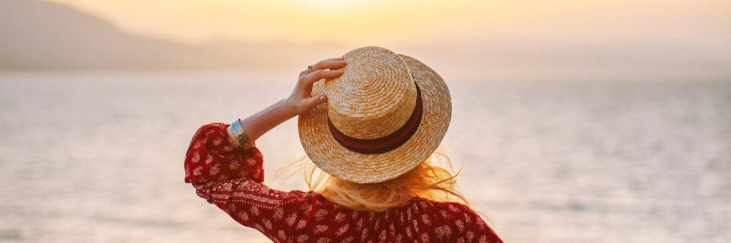 woman stands on beach hand on hat facing backward watching awesome sunset