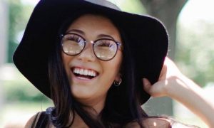 woman wearing eyelash happily smiles stands holding black hat in park