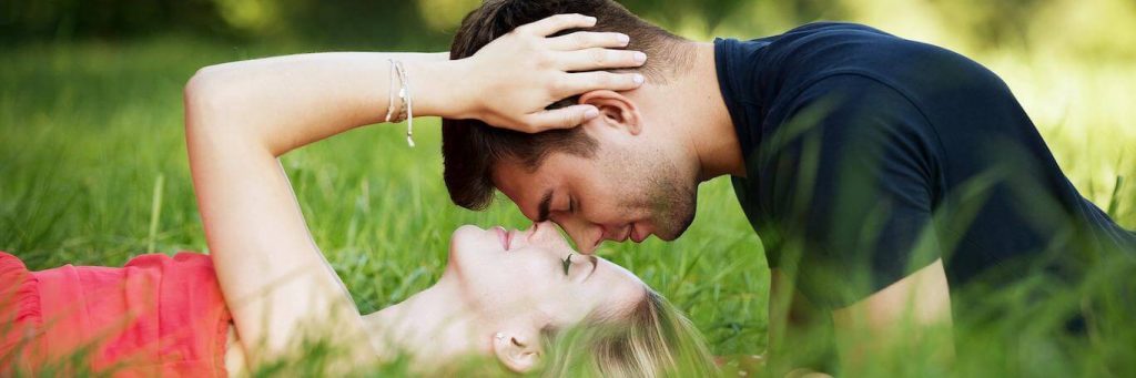 woman lies on green grass while partner sitting kissing