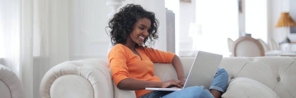 curly black hair woman sits on white couch smiles works on laptop in nice cozy living room