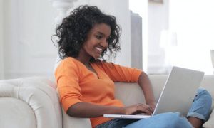 curly black hair woman sits on white couch smiles works on laptop in nice cozy living room