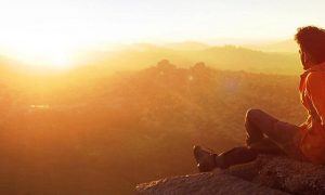 man sits on mountain top wearing back bag watching sunset