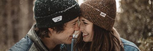 couple wearing winter hat stands smiling in sunny snowy sky