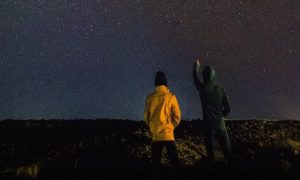 couple stands on rock watching dark blue sky while man pointing at stars