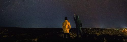 couple stands on rock watching dark blue sky while man pointing at stars
