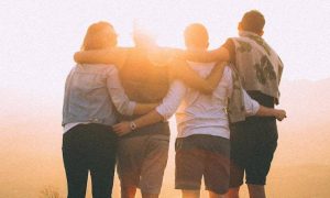 four people stands facing backward hugging watching sunset