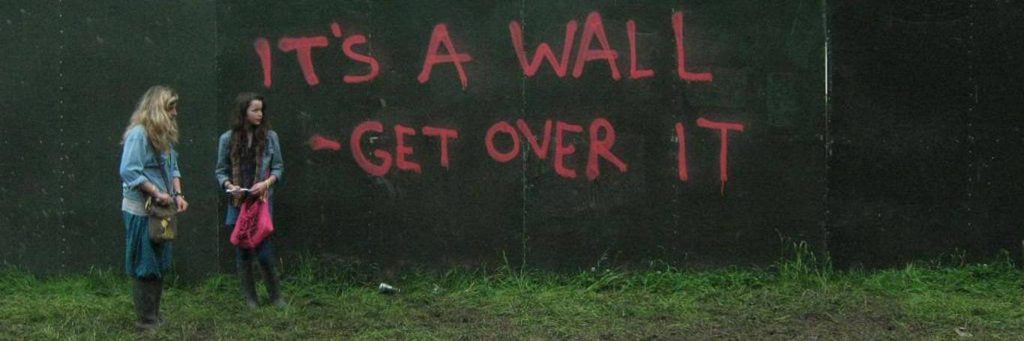 women stand beside black wall pink graffiti sentence saying its a wall get over it