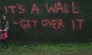 women stand beside black wall pink graffiti sentence saying its a wall get over it