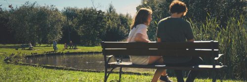 couple sits on bench opposite to lake in park talking