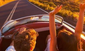 couple sits in car driving on empty road while woman excitedly raising hands in beautiful sunny sky