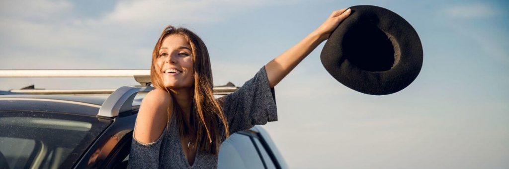 woman stands raising hand with black hat from car window happily smiling