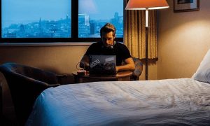 man sits beside bed in bedroom working on laptop on side table at late night