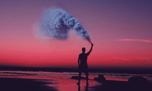 man stands on beach spraying blue gas in sunset sky